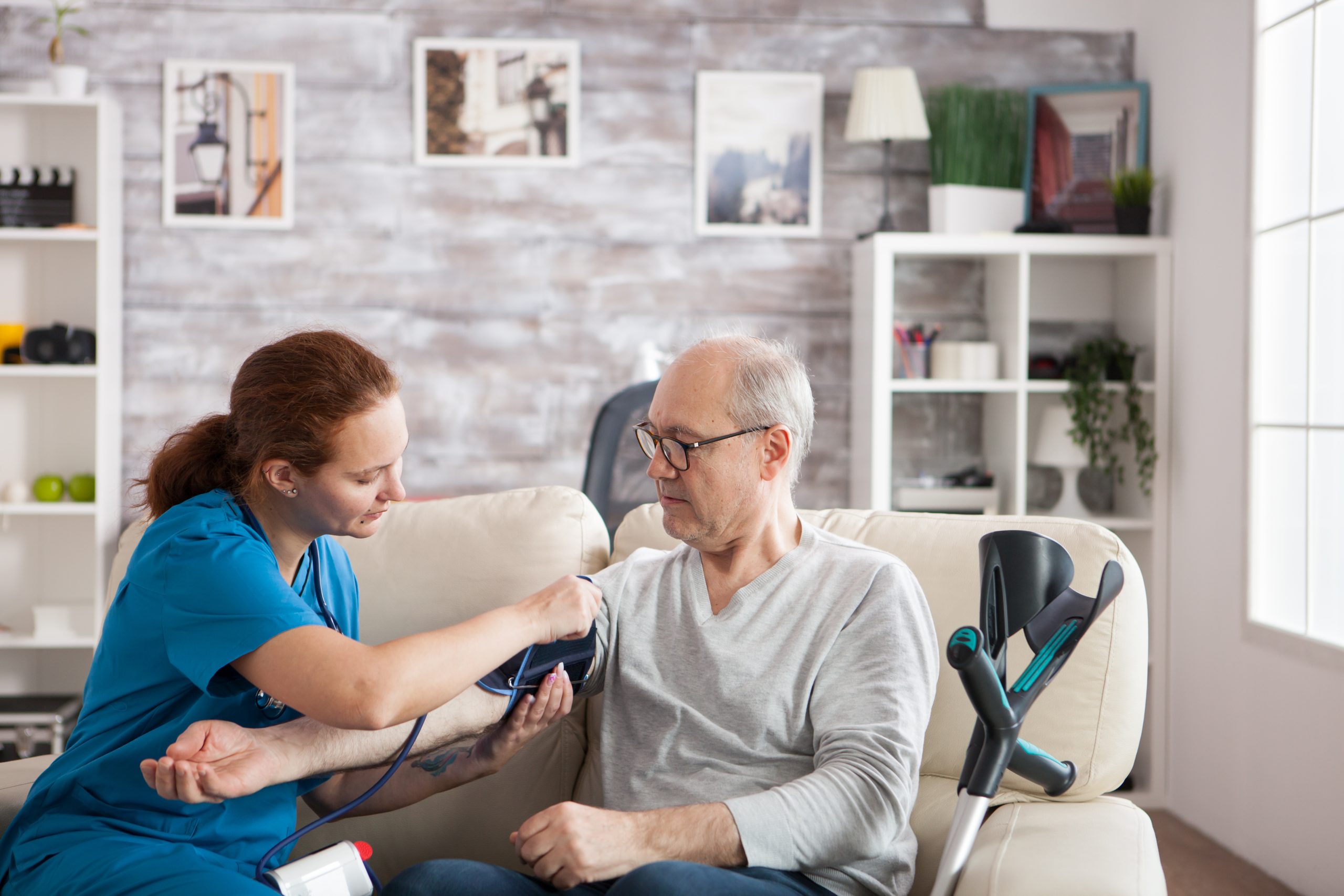 A healthcare worker measures an older man's blood pressure as he sits on a couch with crutches beside him in a living room setting.