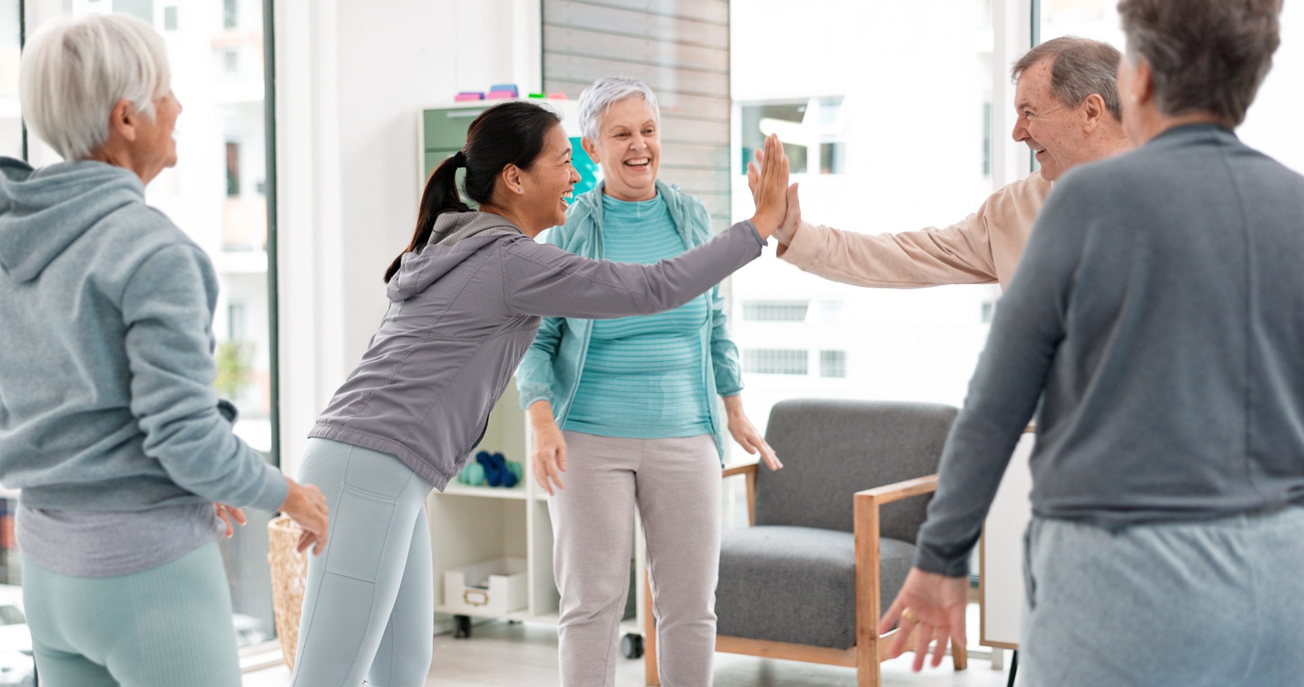 A group of older adults in casual activewear stand in a bright room, smiling and giving high-fives during a group activity or exercise session.